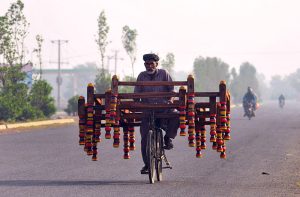 An elderly vendor transports wooden charpai frames on a bicycle along Vehari Road while going about his daily livelihood.