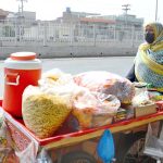A woman pushes a handcart loaded with snack items for sale along a city road to earn livelihood