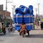 A man transports a huge load of plastic water tanks on a motorcycle rickshaw along a busy city road, posing risk to traffic flow