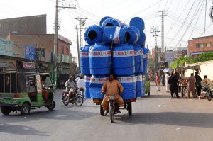 A man transports a huge load of plastic water tanks on a motorcycle rickshaw along a busy city road, posing risk to traffic flow