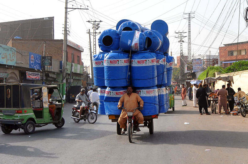 A man transports a huge load of plastic water tanks on a motorcycle rickshaw along a busy city road, posing risk to traffic flow