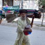 An elderly vendor carries household cleaning tools on his shoulder and walks along the roadside, offering them for sale