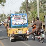 A motorcyclist uses his leg to push a broken-down rickshaw towards a nearby repair workshop along a city road