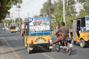 A motorcyclist uses his leg to push a broken-down rickshaw towards a nearby repair workshop along a city road