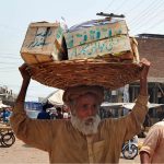 An elderly laborer carries a heavy basket loaded with wooden crates on his head while passing through a bustling wholesale market, earning his livelihood amid the scorching summer heat in the city