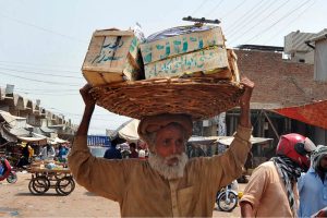 An elderly laborer carries a heavy basket loaded with wooden crates on his head while passing through a bustling wholesale market, earning his livelihood amid the scorching summer heat in the city