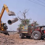 Workers use heavy machinery to load a concrete sewer pipe onto a tractor trolley during ongoing sewerage development work aimed at improving drainage infrastructure in the city