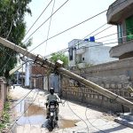 A motorcyclist navigates beneath a storm-damaged electricity pole that leans dangerously across a residential street with tangled electric wires hanging low over the road and posing a serious threat to residents, as authorities have yet to clear the hazard