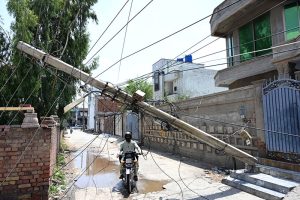 A motorcyclist navigates beneath a storm-damaged electricity pole that leans dangerously across a residential street with tangled electric wires hanging low over the road and posing a serious threat to residents, as authorities have yet to clear the hazard