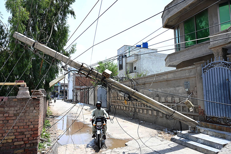 A motorcyclist navigates beneath a storm-damaged electricity pole that leans dangerously across a residential street with tangled electric wires hanging low over the road and posing a serious threat to residents, as authorities have yet to clear the hazard