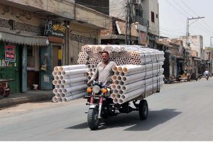 A transporter carries a large load of PVC pipes on a motorcycle loader while passing through a city street, delivering construction materials for ongoing development work.