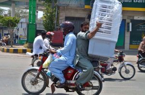 A person sitting on rear seat of motorbike holding a room cooler in a dangerous way heading towards destinantion at Khayyam chowk.
