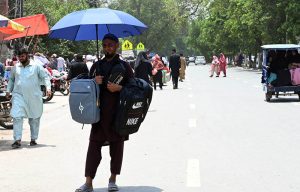 A street vendor sells school bags on a roadside stall, using an umbrella for shade while enduring the intense sun in the city.