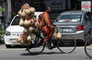 A vendor cycles through a busy road carrying a large collection of handcrafted gourd bird feeders for sale.