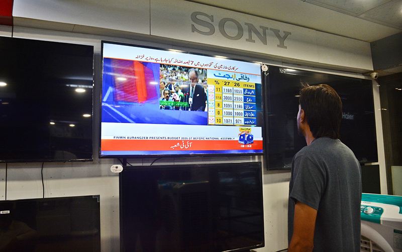 A shopkeeper keeps his eyes fixed on a television screen at his shop while watching Finance Minister Muhammad Aurangzeb present the Federal Budget 2026–27