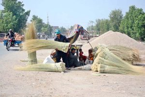 A roadside vendor arranges bundles of brooms for sale while his family sits under a temporary shade to escape the scorching sun beside their roadside stall in the city.