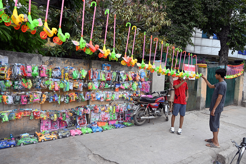Children browse through a colourful display of plastic toys including toy cars, push toys and various playthings at a roadside toy stall