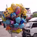 A young boy sells colourful foil balloons featuring popular cartoon characters including Doraemon, monkeys, sharks, bears and stars