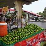 A young boy operates a fresh lemon juice pressing cart loaded with green limes at a busy roadside stall