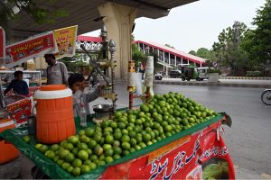 A young boy operates a fresh lemon juice pressing cart loaded with green limes at a busy roadside stall