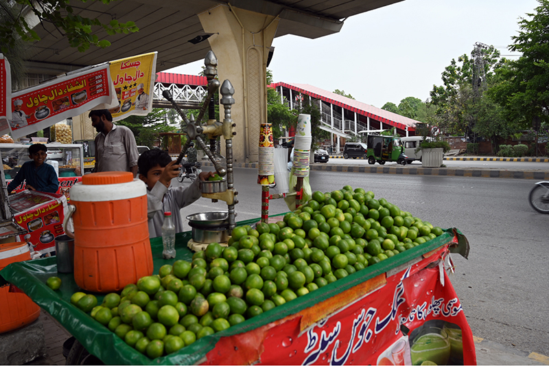 A young boy operates a fresh lemon juice pressing cart loaded with green limes at a busy roadside stall