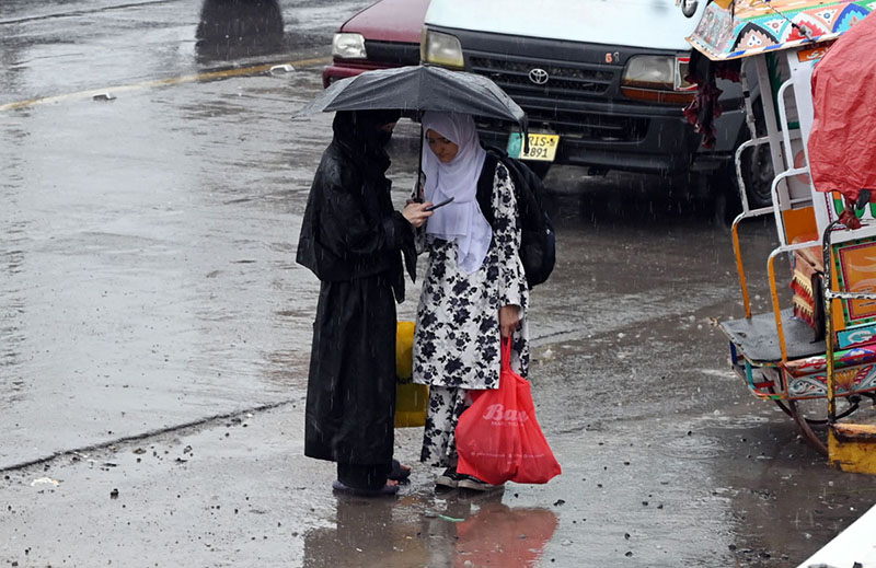Girls share an umbrella while waiting for a bus on a waterlogged as monsoon rains lash in the Federal Capital