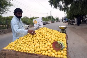 A young boy operates a fresh lemon juice pressing cart loaded with green limes at a busy roadside stall