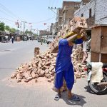 A laborer carries a heavy bundle of bricks on his back during the urban development despite the sweltering summer heat at a construction site in Sarfaraz Colony