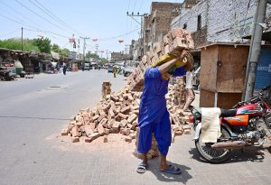 A laborer carries a heavy bundle of bricks on his back during the urban development despite the sweltering summer heat at a construction site in Sarfaraz Colony