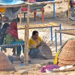 A nomadic woman weaves traditional baskets from dried tree branches under a makeshift roadside shelter to earn a living, while her family rests in a nearby temporary hut amidst the intense heat