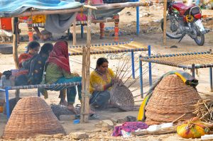 A nomadic woman weaves traditional baskets from dried tree branches under a makeshift roadside shelter to earn a living, while her family rests in a nearby temporary hut amidst the intense heat