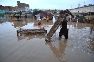 A man carries his household belongings to a safer area after heavy monsoon rains inundated a makeshift settlement, forcing families to relocate from flood-affected homes.