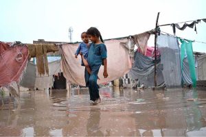 A man carries his household belongings to a safer area after heavy monsoon rains inundated a makeshift settlement, forcing families to relocate from flood-affected homes.
