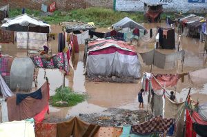 A man carries his household belongings to a safer area after heavy monsoon rains inundated a makeshift settlement, forcing families to relocate from flood-affected homes.