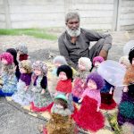 An elderly vendor displays colorful toy dolls for sale on his roadside setup, hoping to attract customers