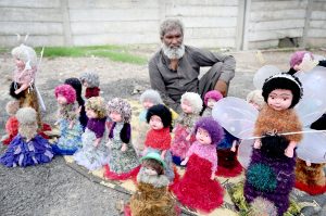 An elderly vendor displays colorful toy dolls for sale on his roadside setup, hoping to attract customers