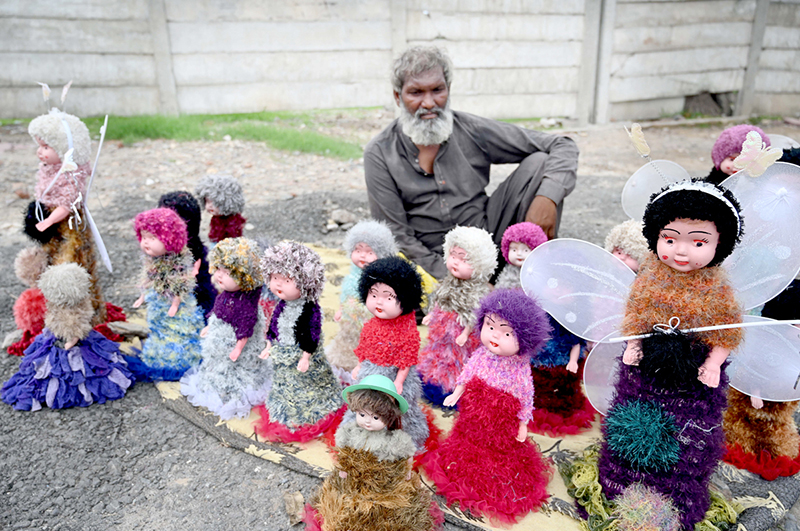 An elderly vendor displays colorful toy dolls for sale on his roadside setup, hoping to attract customers