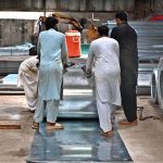 Workers are manually unrolling a heavy steel coil to make door frames (chokhat) in a warehouse on City Saddar Road