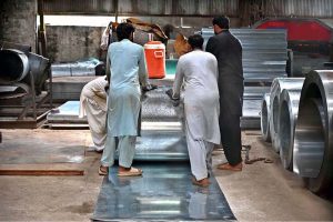 Workers are manually unrolling a heavy steel coil to make door frames (chokhat) in a warehouse on City Saddar Road
