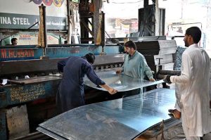 Workers are manually unrolling a heavy steel coil to make door frames (chokhat) in a warehouse on City Saddar Road