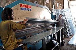 Workers are manually unrolling a heavy steel coil to make door frames (chokhat) in a warehouse on City Saddar Road