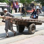 An elderly man pulls a handcart loaded with bamboo poles through a street