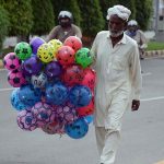 An elderly street vendor carries inflatable handballs while strolling through a street in search of customers