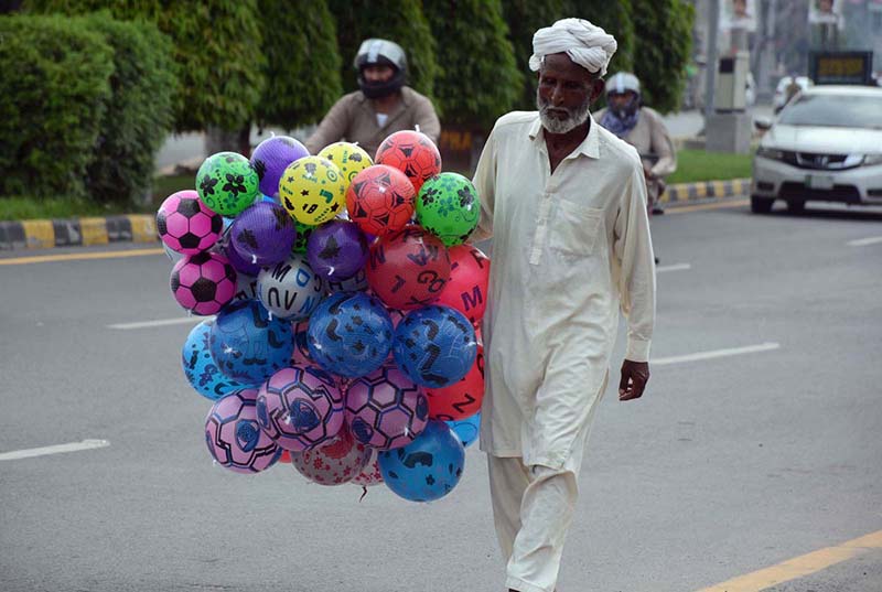 An elderly street vendor carries inflatable handballs while strolling through a street in search of customers