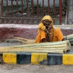 An elderly woman vendor sits beside bundles of brooms displayed for sale along a roadside, waiting for customers to earn her livelihood in the city