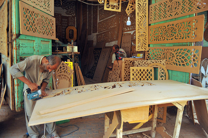 An artisan skillfully shapes a wooden panel with a jigsaw while crafting intricate traditional jali work at a local workshop