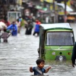 A child stands amid floodwater as an auto-rickshaw makes its way through the inundated street following heavy monsoon rains in Dhoke Khabba