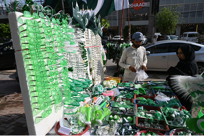A vendor displays Pakistani flags, badges, headbands and other Jashn-e-Azadi items at a roadside stall near Commercial Market as shoppers browse the festive merchandise ahead of Independence Day celebrations