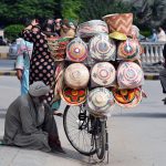 A vendor sits beside his bicycle loaded with colourful handwoven baskets, displayed for sale