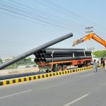 Labourers use an excavator to unload large plastic pipes from a truck along a road during ongoing development work